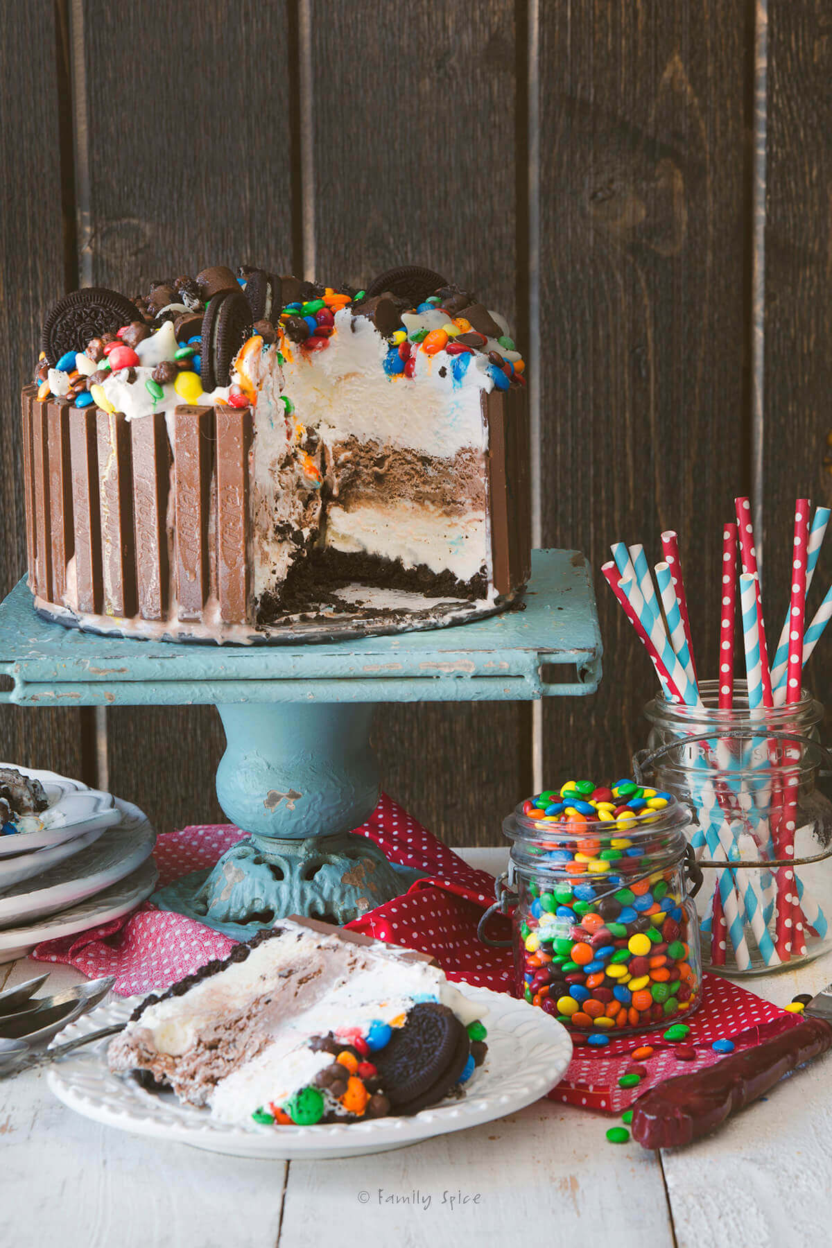 Ice cream cake surrounded by Kit Kats and topped with candies and cookies on a cake stand with a slice on a plate next to it