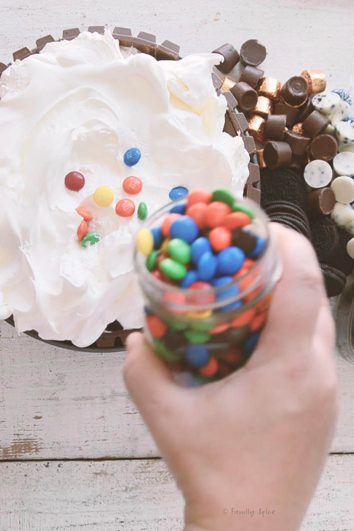 Mini M&Ms in a small mason jar being poured over the top of an ice cream cake topped with whipped cream