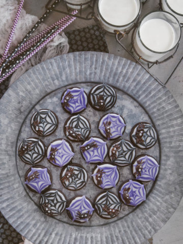 Overhead view of raisin spider cookies on a rustic metal tray with glasses of milk and black and purple straws