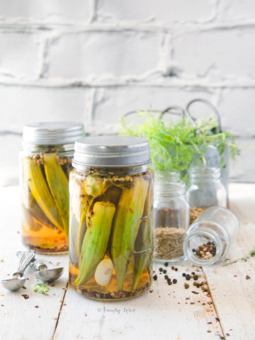 Two jars of pickled okra on a white background with fresh herbs and ingredients behind it