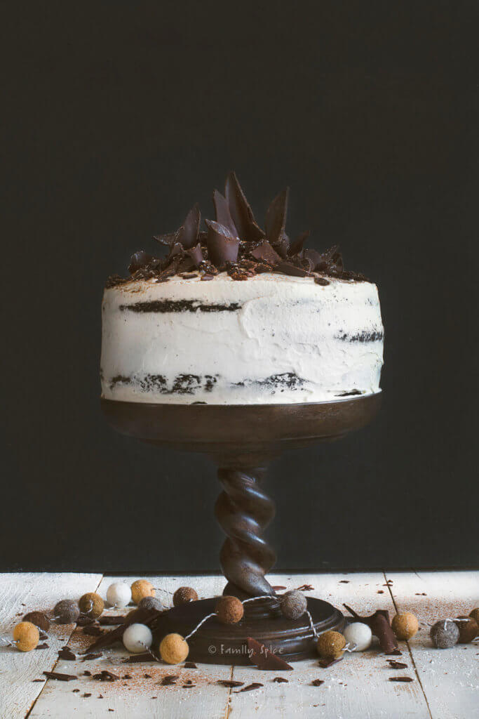 A whipped cream frosted raisin black forest cake with chocolate shards on top on a dark brown cake pedestal
