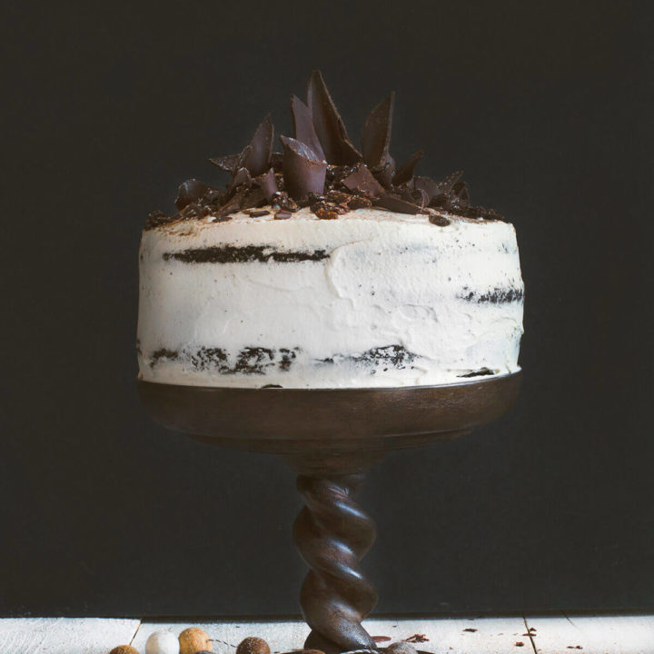 A whipped cream frosted raisin black forest cake with chocolate shards on top on a dark brown cake pedestal