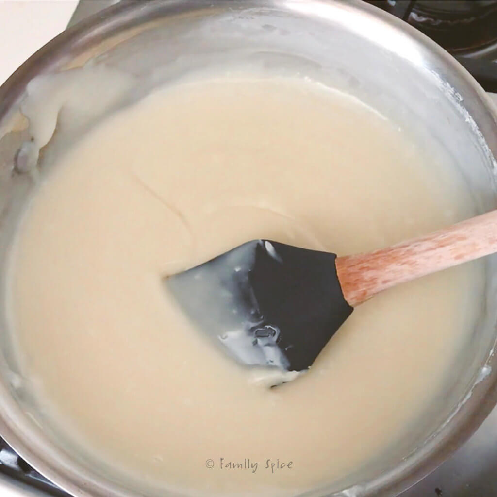 Melted white chocolate chips mixed with sweetened condensed milk in a stainless pot with a rubber spatula in it