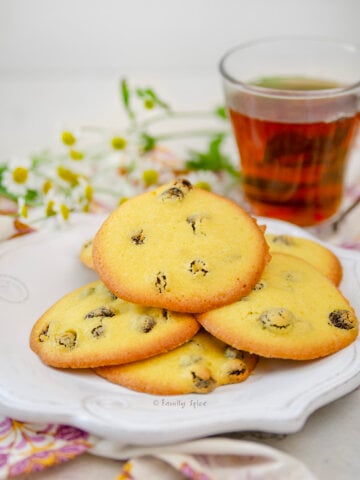 A white plate with Persian Saffron Raisin Cookies (shirini kishmishi) on it, plus flowers and cup of tea behind it.