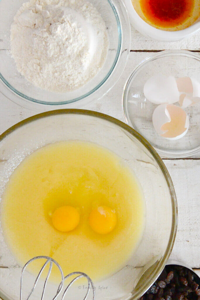 A large mixing bowl with eggs added to a melted butter mixture with other bowls with ingredients next to it