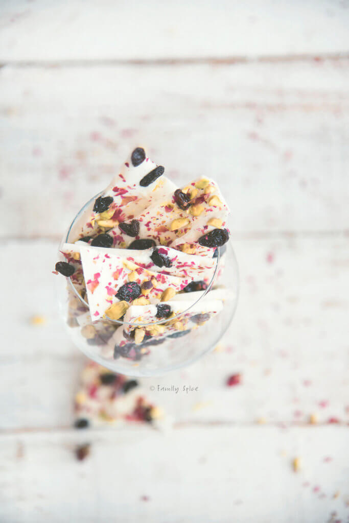 Top view of white chocolate bark topped with pisatchios, raisins and rose petals broken up and in a wine glass