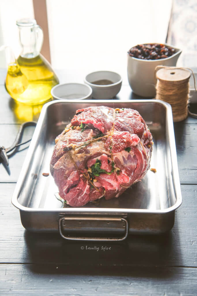 A boneless and stuffed leg of lamb rolled and tied with kitchen twine in a roasting pan with ingredients around it