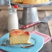 Closeup of a slice of pineapple upside down cake on blue plate with milk and the rest of the cake behind it