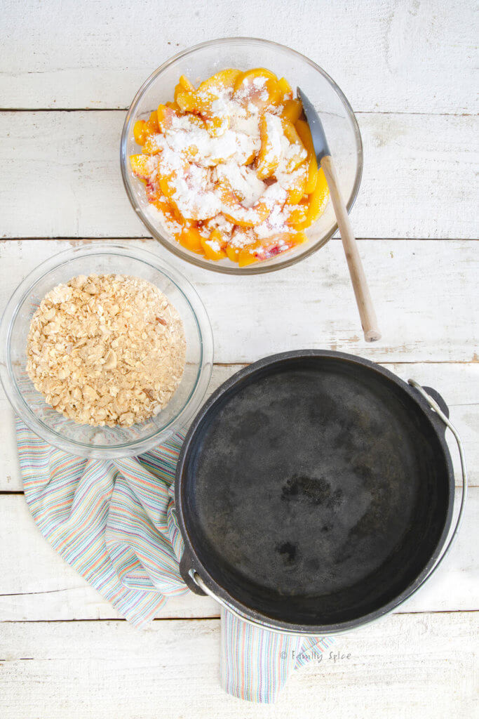 Top view of an empty Dutch oven with a bowl of sliced peaches topped with flour and another bowl with crisp ingredients mixed in