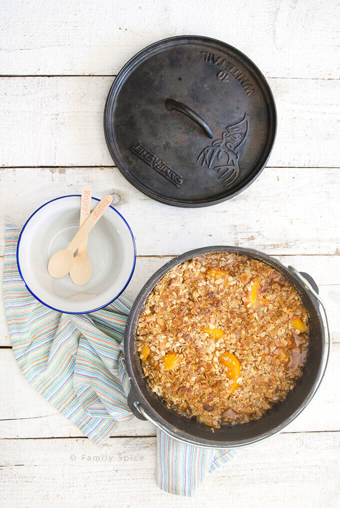 Overhead shot of freshly baked Dutch oven peach crisp with almonds with empty bowls by FamilySpice.com