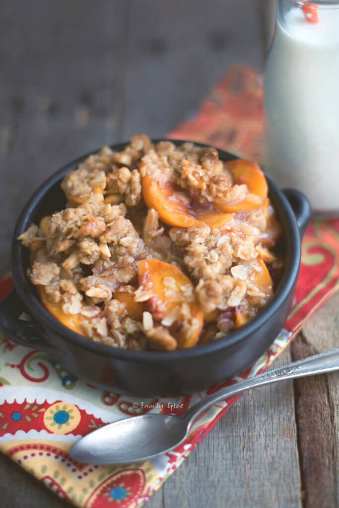 Closeup of a dark bowl with peach crisp in it and a milk bottle next to it