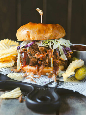 A stuffed pulled pork sandwich dripping with barbecue sauce and served with chips and pickles