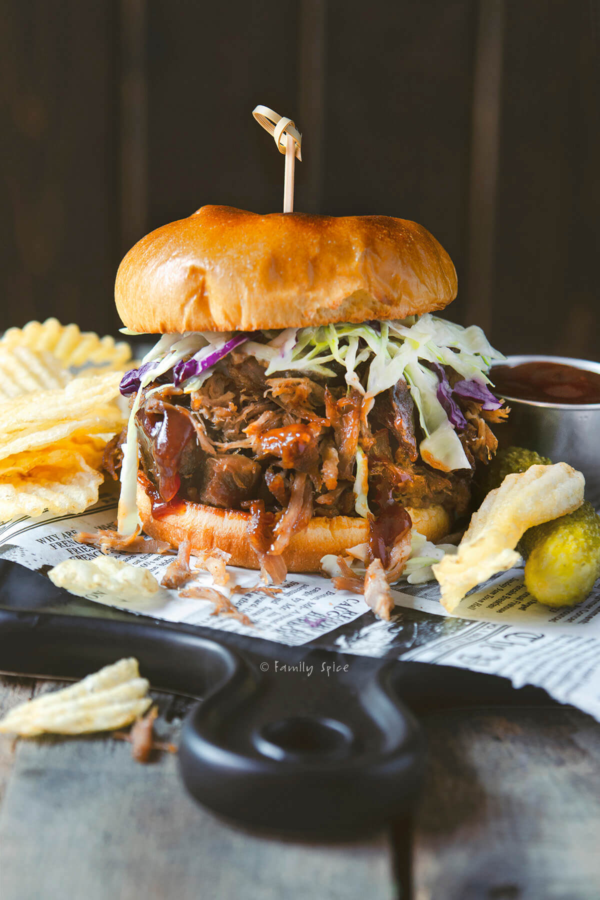 A stuffed pulled pork sandwich dripping with barbecue sauce and served with chips and pickles