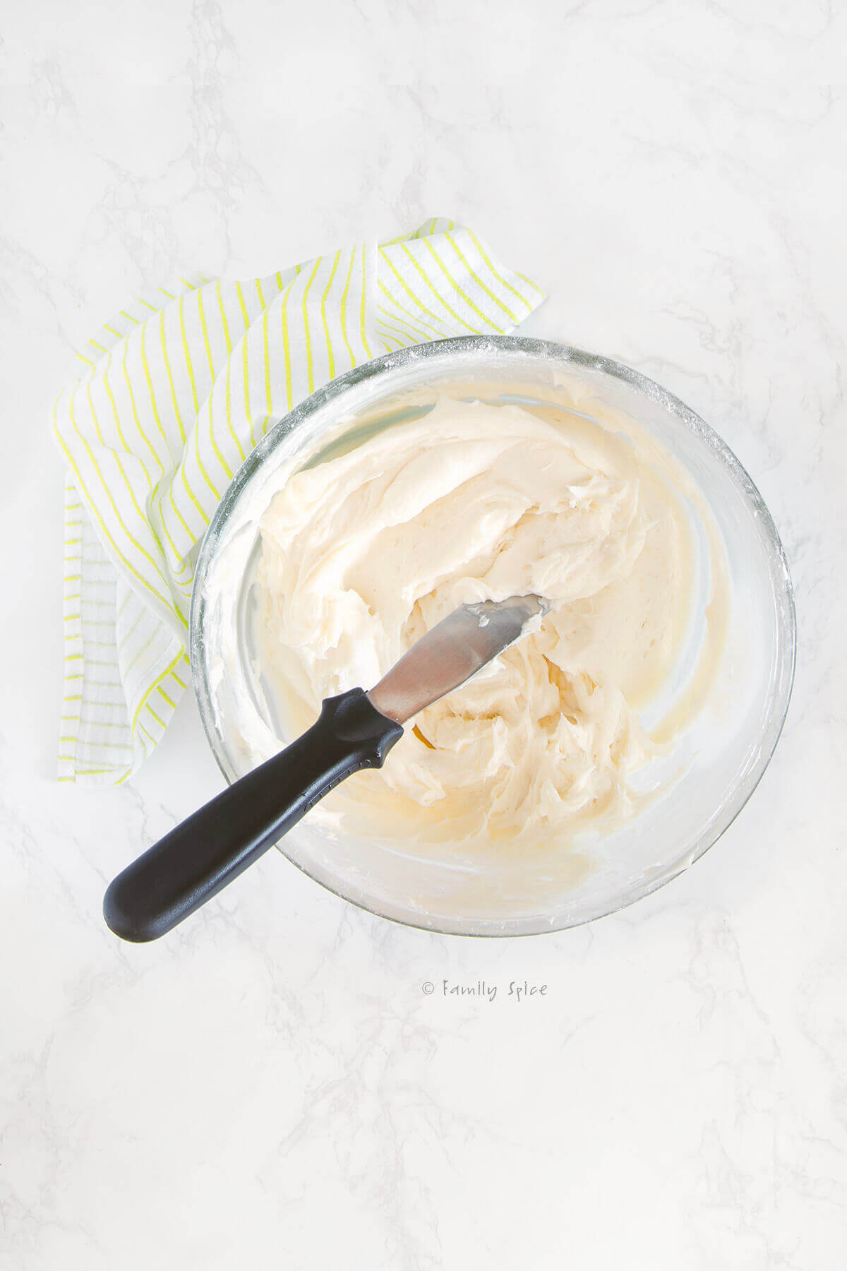 A glass mixing bowl with cream cheese frosting and a metal spreader