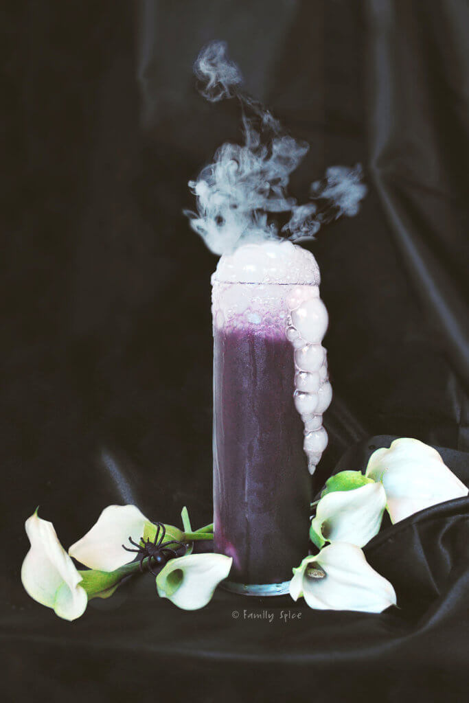A tall glass of purple halloween drink with bubbles and smoke coming out of it