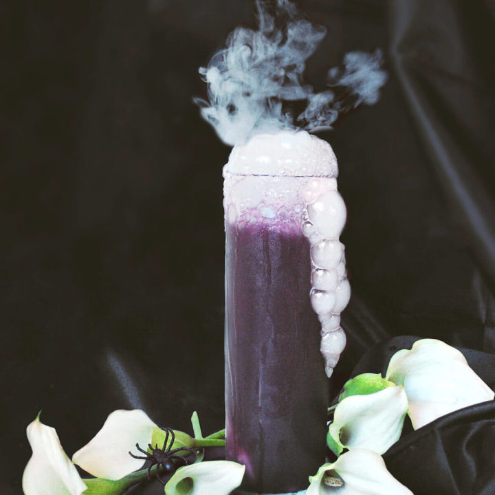 A tall glass of purple halloween drink with bubbles and smoke coming out of it