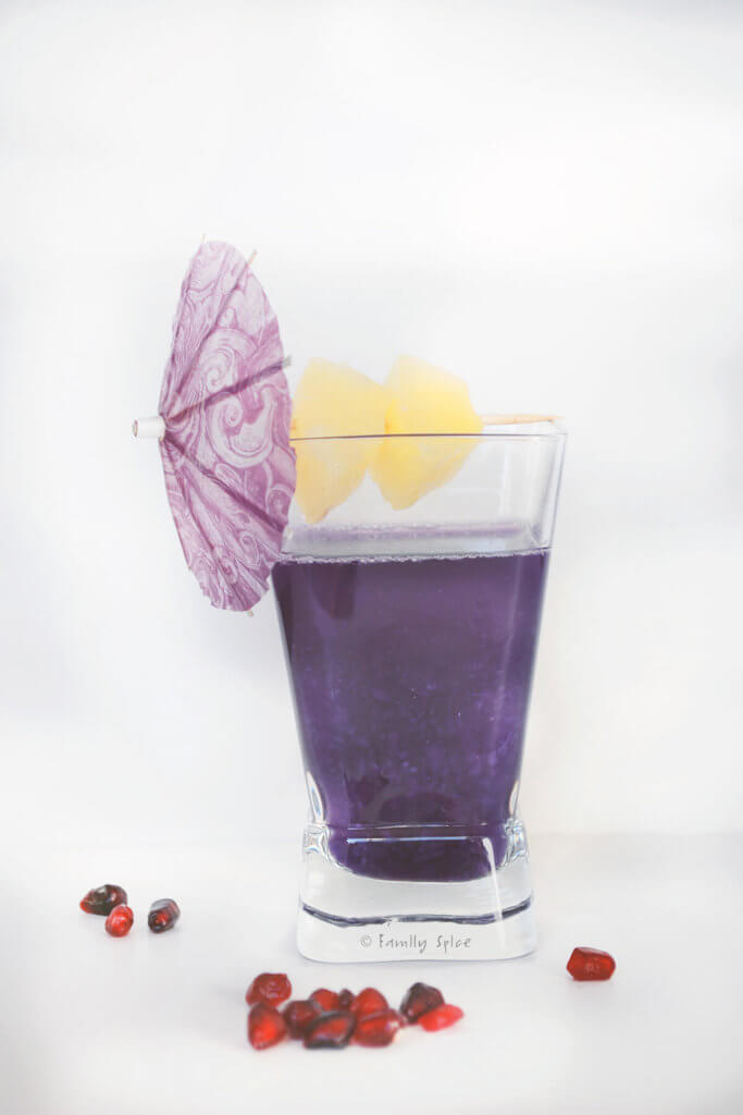 A small glass with purple cocktail, garnished with pineapple and purple umbrella with some pomegranate arils around it