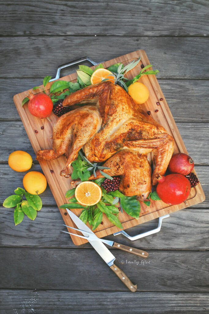 Overhead image of butterflied turkey with oranges, pomegranates and fresh herbs on a cuttingboard with carving knife and fork