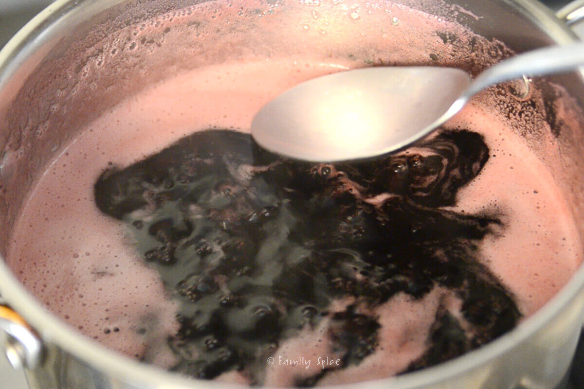 A spoon removing foam from top of pomegranate jelly mixture in a stainless steel pot