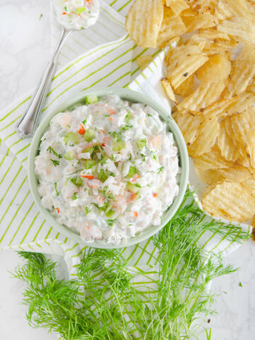 Closeup of top view of a bowl with shrimp and crab dip with a spoon, fresh dill and potato chips around it