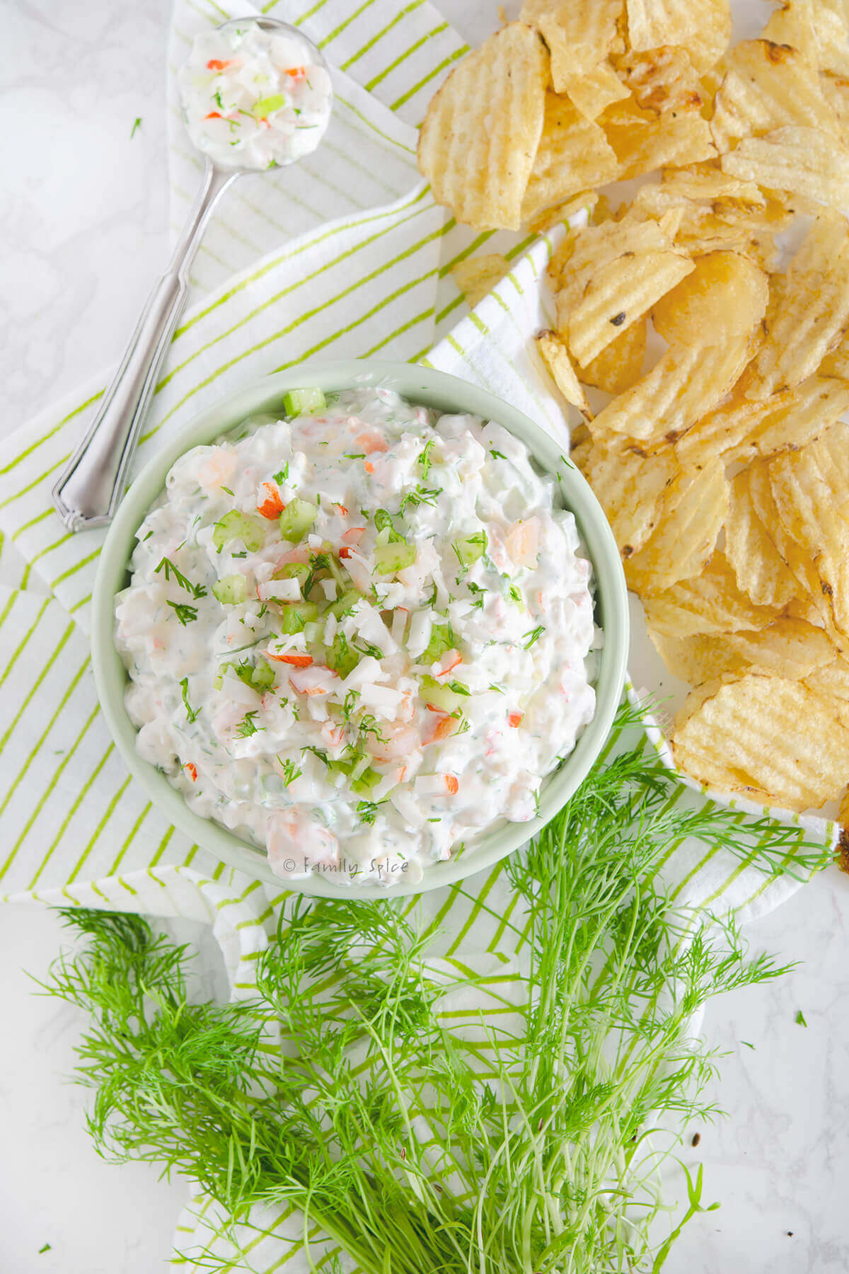Closeup of top view of a bowl with shrimp and crab dip with a spoon, fresh dill and potato chips around it