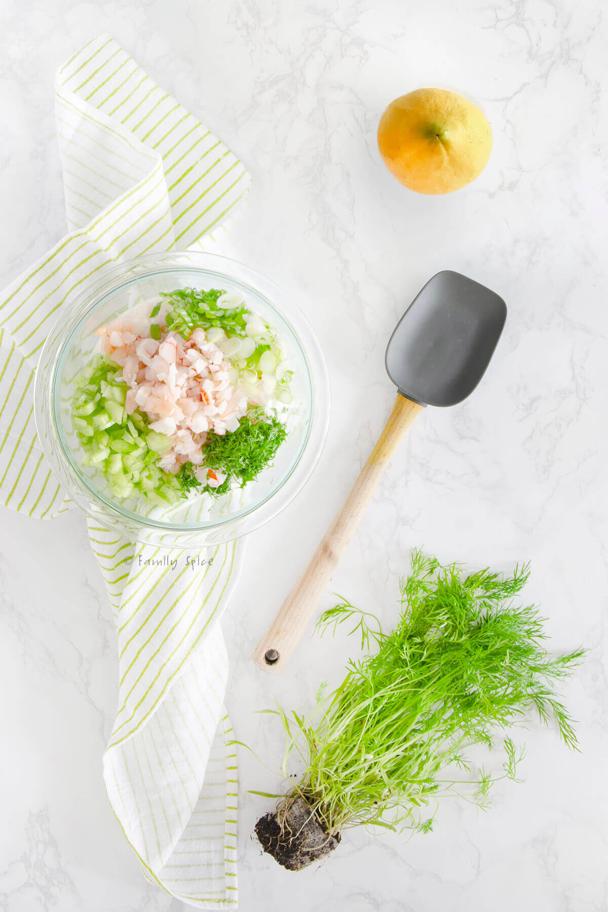 Ingredients to make shrimp and crab dip in a glass bowl with a lemon, dill plant and rubber spatula next to it