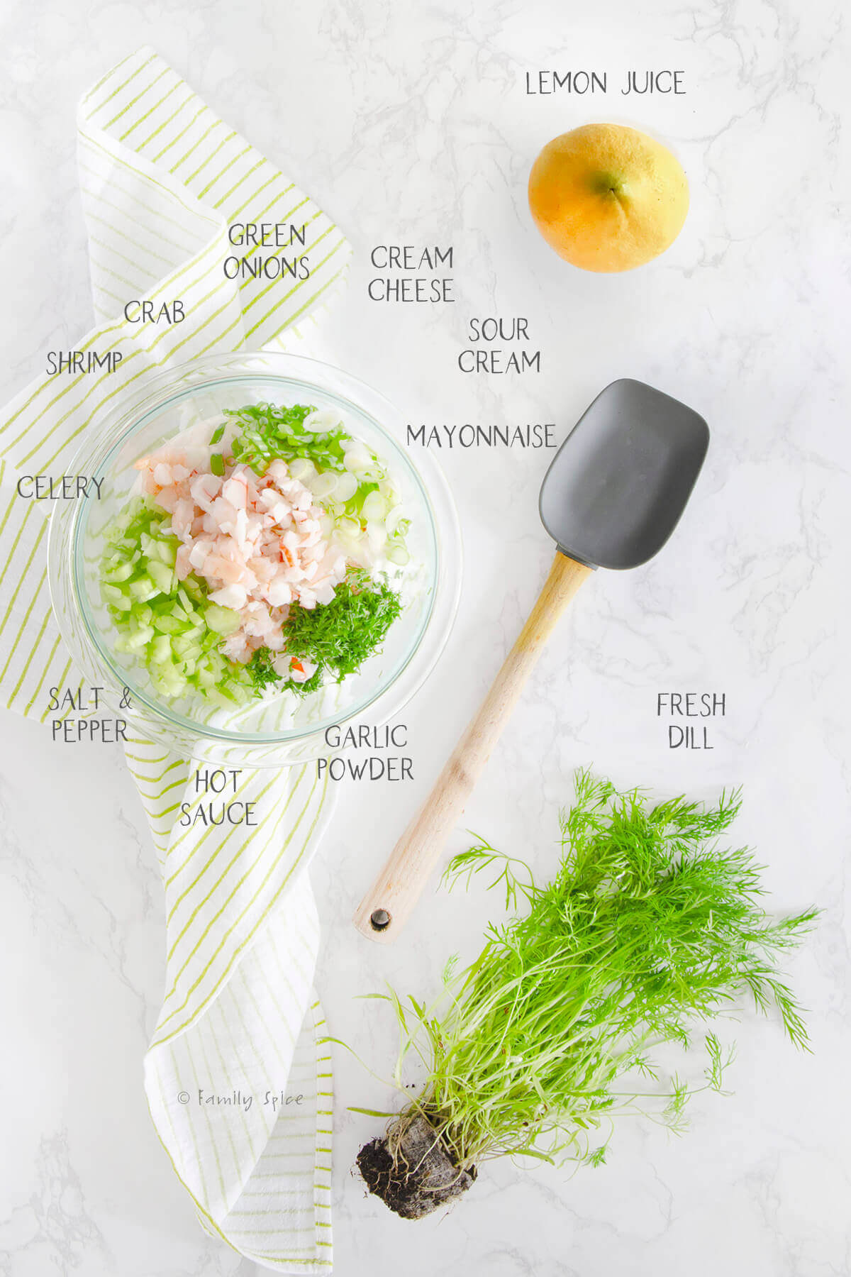 Ingredients needed and labeled to make shrimp and crab dip in a glass bowl with a lemon, dill plant and rubber spatula next to it