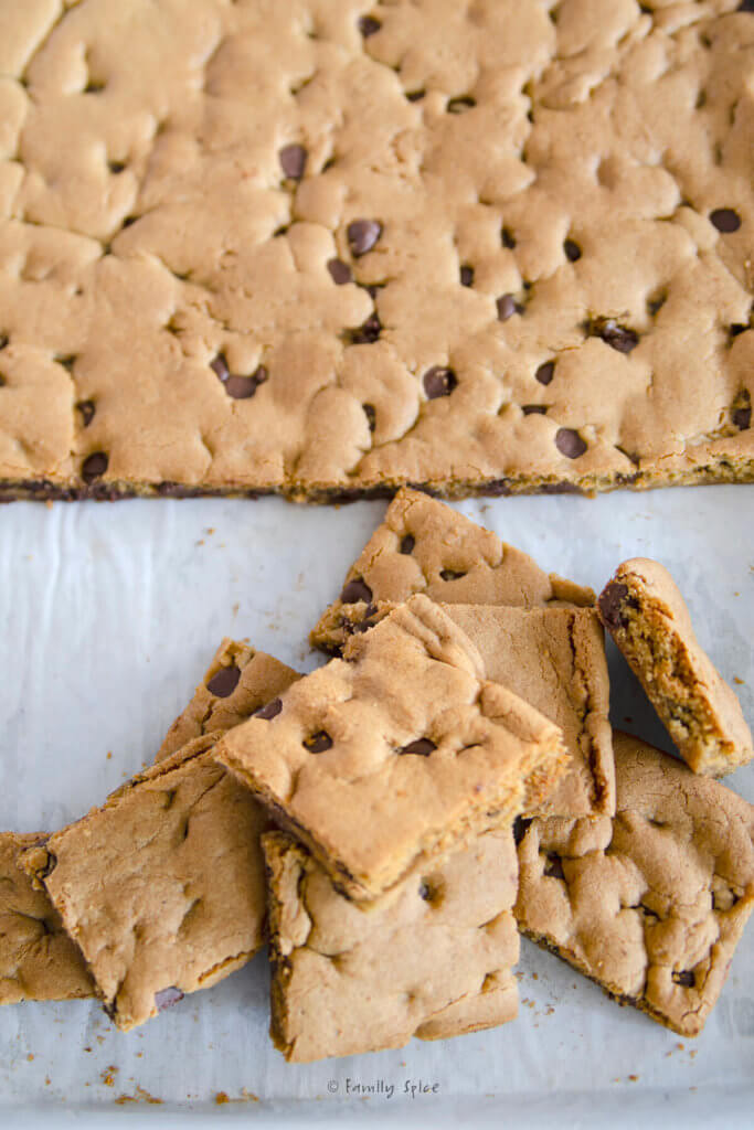 A small pile of chocolate chip cookie bars on a sheet pan with the uncut bars behind it