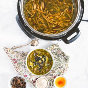 A bowl of ash reshteh with small bowls holding toppings and the instant pot with more soup in it