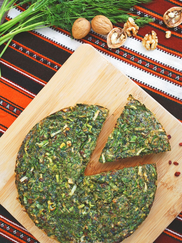 Kuku sabzi on a wood cuttingboard with a slice cut out and herbs and nuts around it