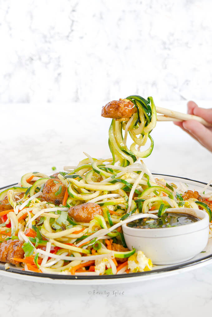 Side view of chopsticks holding a piece of orange chicken with Zucchini Noodle Pad Thai by FamilySpice.com
