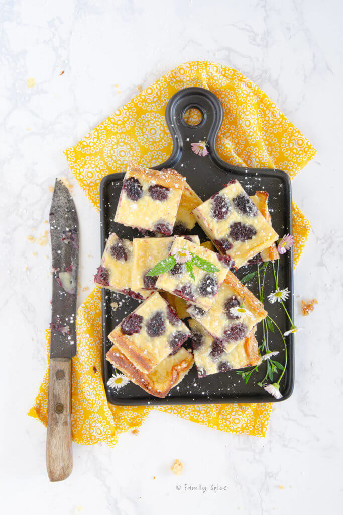 Blackberry custard bars piled up on a black cutting board with yellow napkin and a knife next to it