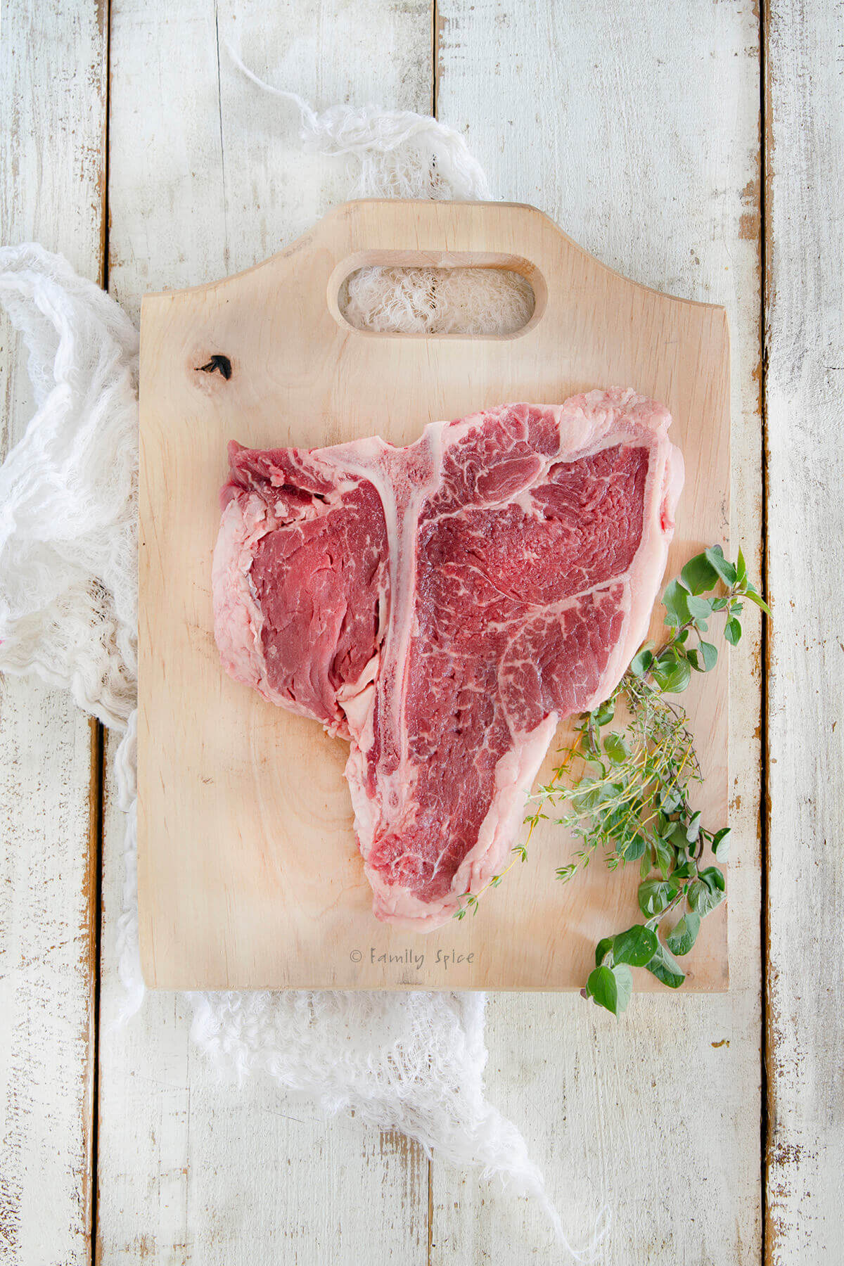 Top view of a raw porterhouse steak on a cutting board with fresh herbs next to it