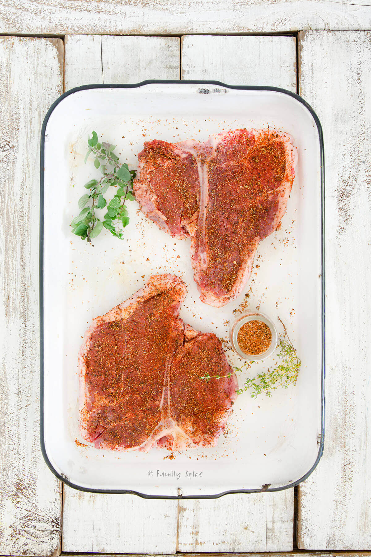 Top view of a white enamel pan with two seasoned raw porterhouse steaks and fresh herbs next to it