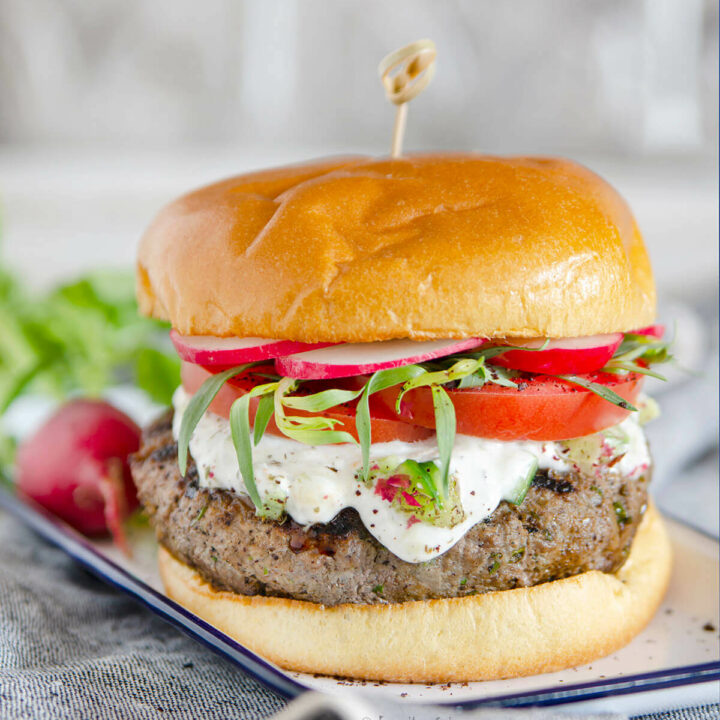 Side view of a kebab burger with yogurt, tomatoes and fresh herbs on a small white enamel plate