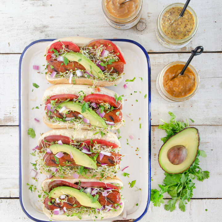 Top view of four gourmet hot dogs with tomatoes, avocado slices and sprouts with a halved avocado and mustard jars next to it