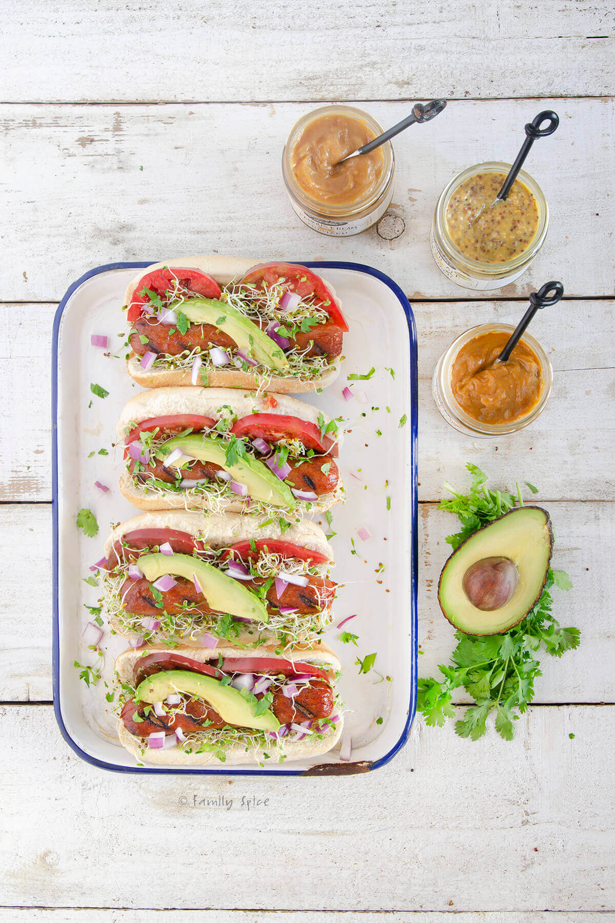 Top view of four gourmet hot dogs with tomatoes, avocado slices and sprouts with a halved avocado and mustard jars next to it
