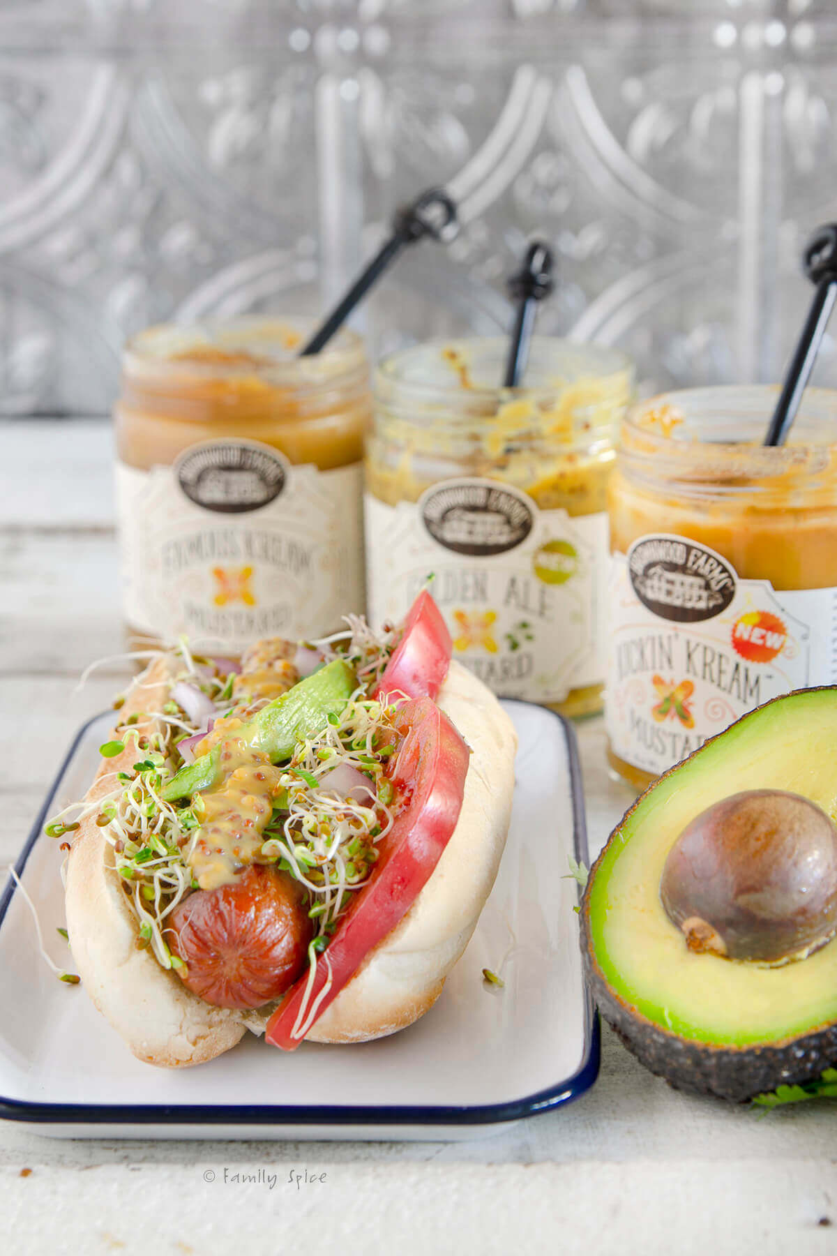 A hot dog with tomatoes, avocado slices and sprouts with a halved avocado and mustard jars behind it