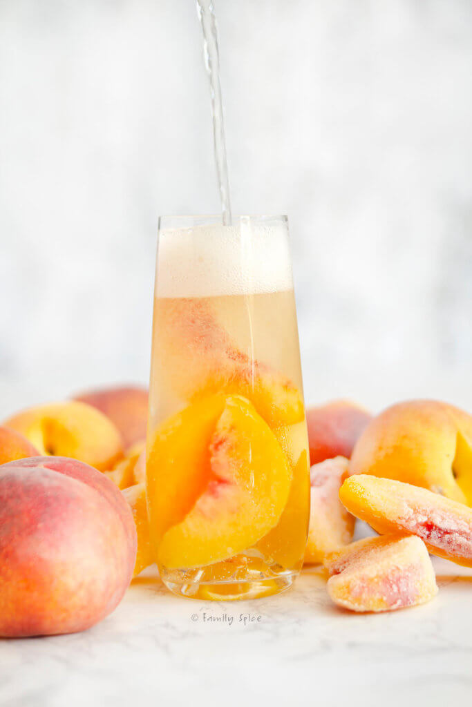 Side view of a tall glass of sparkling peach sangria with more sparkling wine being poured in and surround by whole peaches and slices