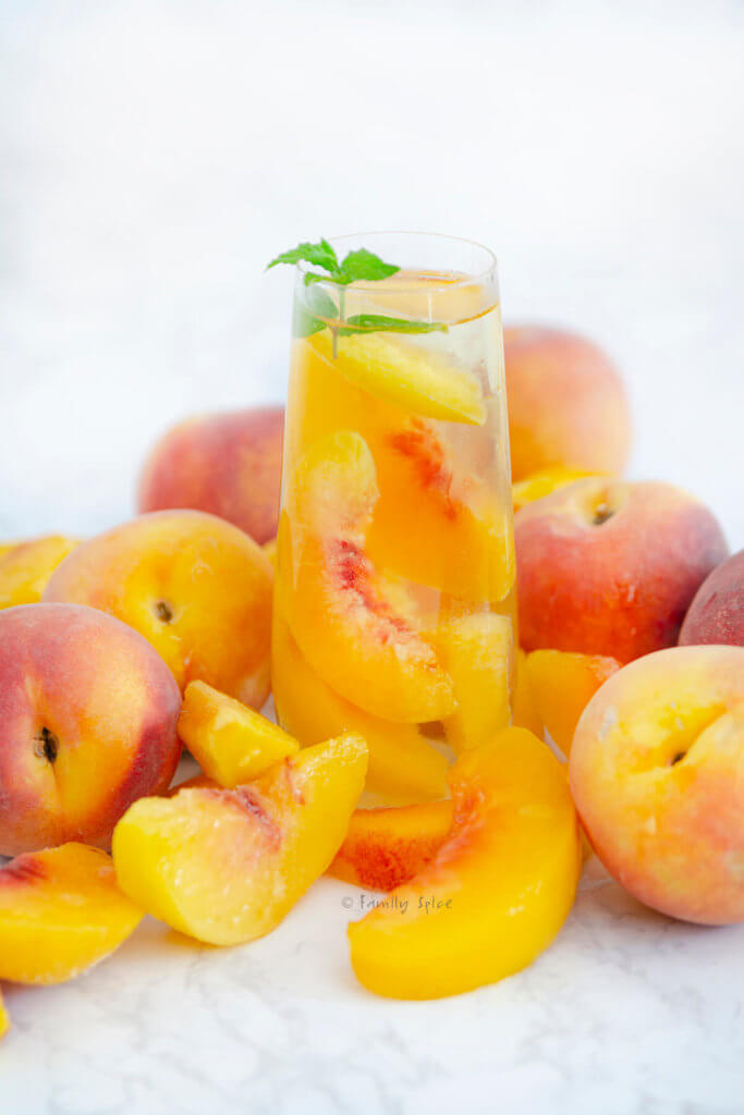 A tall glass of sparkling peach sangria topped with fresh mint surround by whole peaches and slices