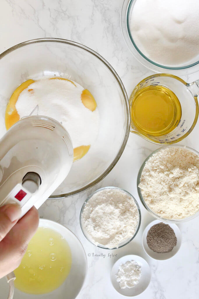 Using a hand blender to mix sugar and egg yolks in a mixing bowl with other ingredients in bowls around it