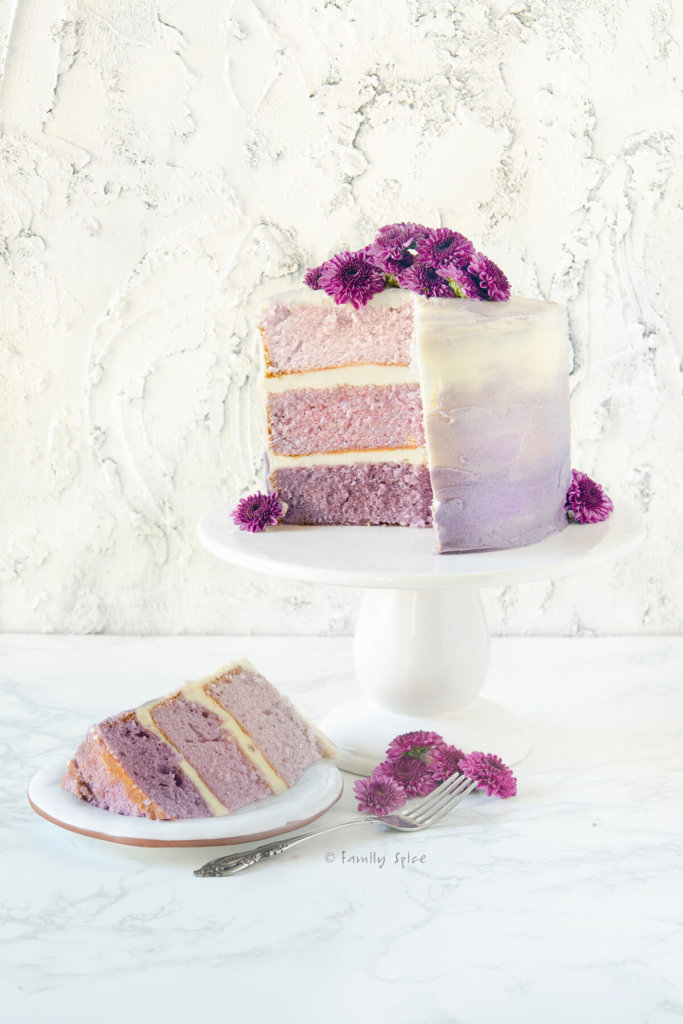 A purple grape kool aid ombre cake with purple flowers on top cut open with a slice on a white plate next to it