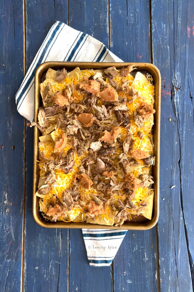 Top view of a baking sheet with tortilla chips, cheese, beans and pulled pork on it