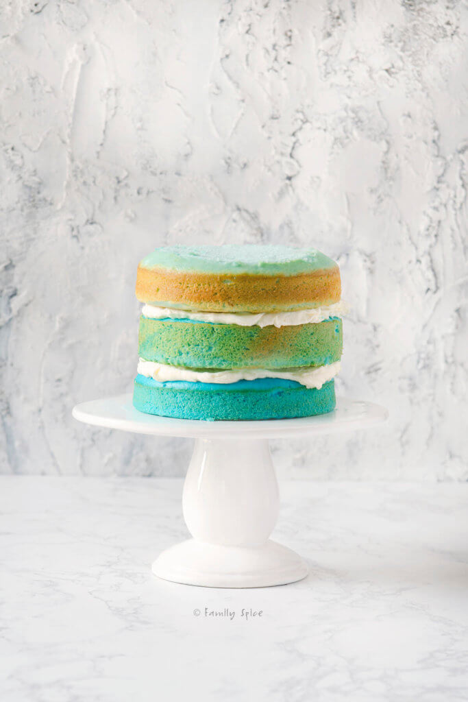 A blue raspberry blue ombre cake not completely frosted on a white cake stand
