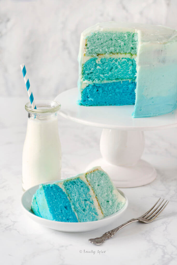 Close up of a blue raspberry blue ombre cake cut open on a white cake stand with a slice on a white plate and a glass bottle of milk next to it