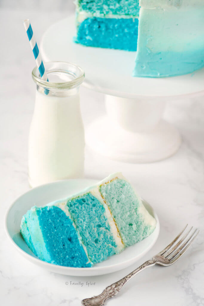 Close up of a slice of blue raspberry kool aid ombre cake with a glass bottle of milk behind it
