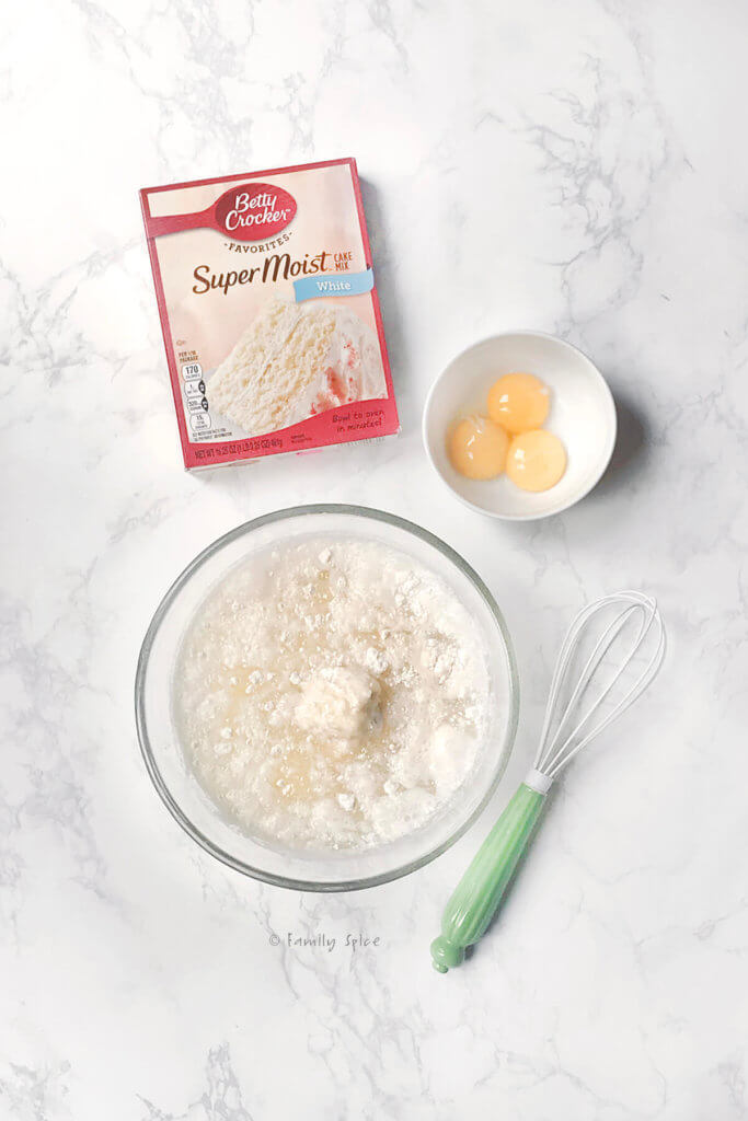 A mixing bowl with white cake mix, a box of white cake mix, a small bowl with egg yolks and a whisk