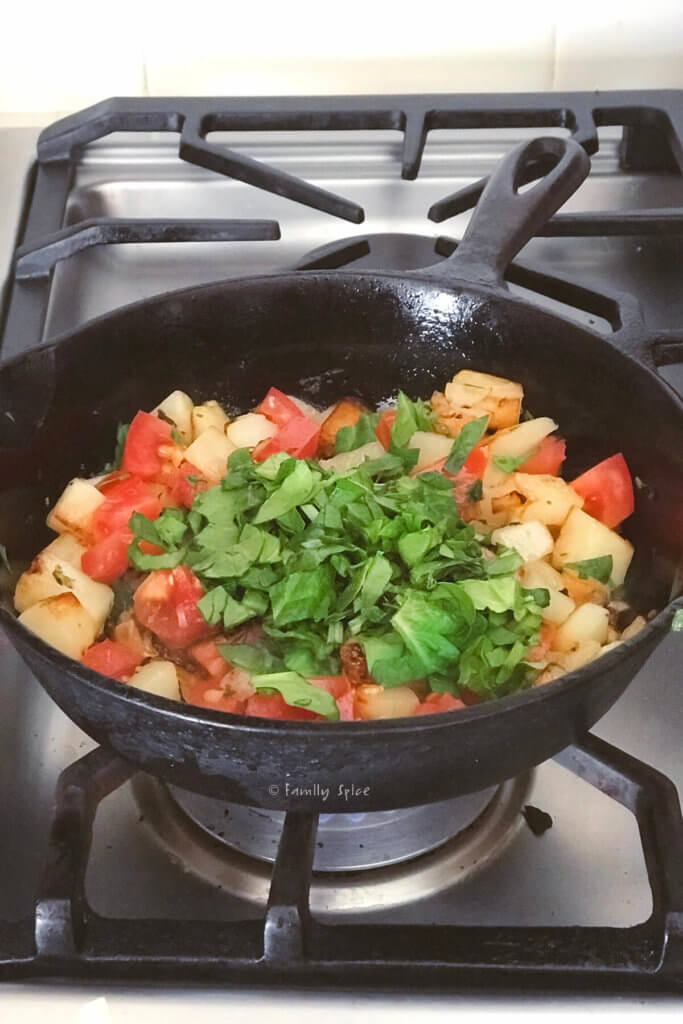 Adding tomatoes and cilantro to cooked potatoes and onions in a cast iron pan