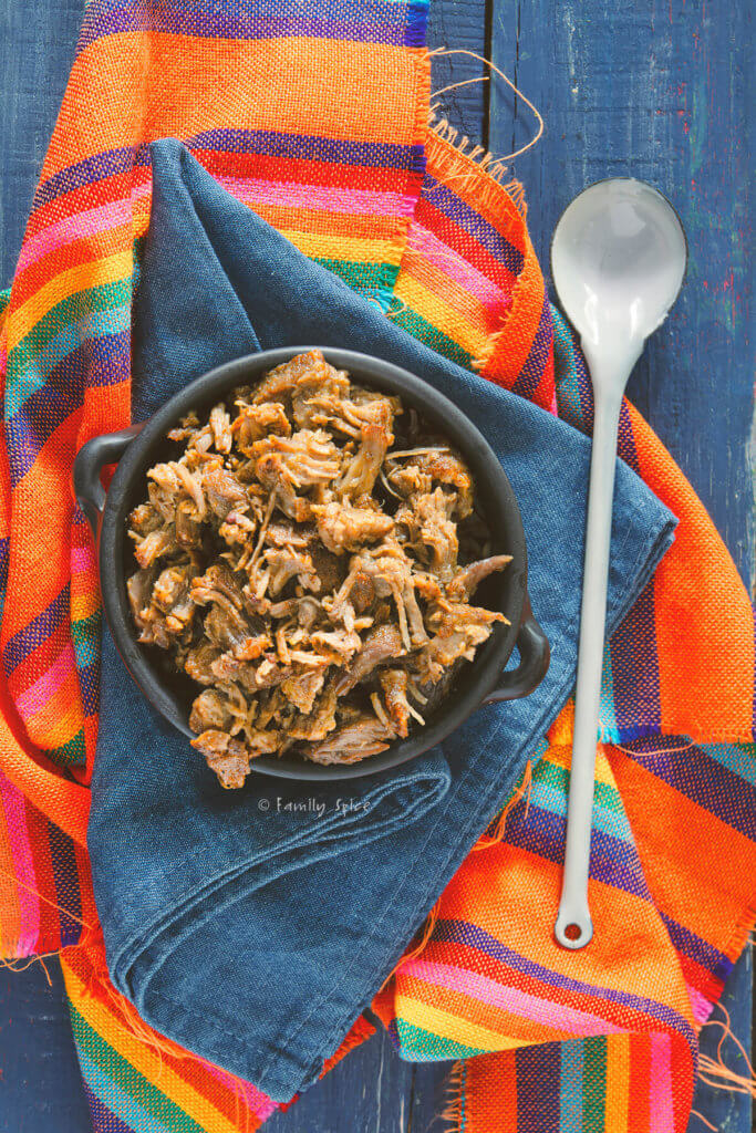 A metal bowl with shredded pork in it on a blue napkin with colorful fabric under it