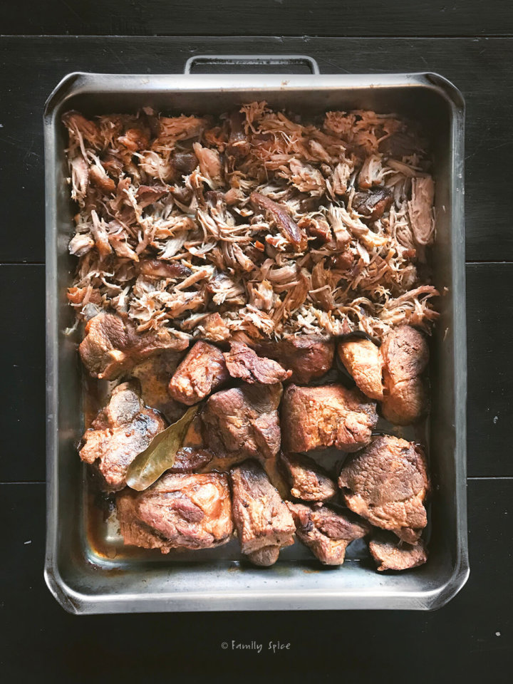 Overhead view of a roasting pan half filled with roasted pork shoulder and half filled with shredded pulled pork